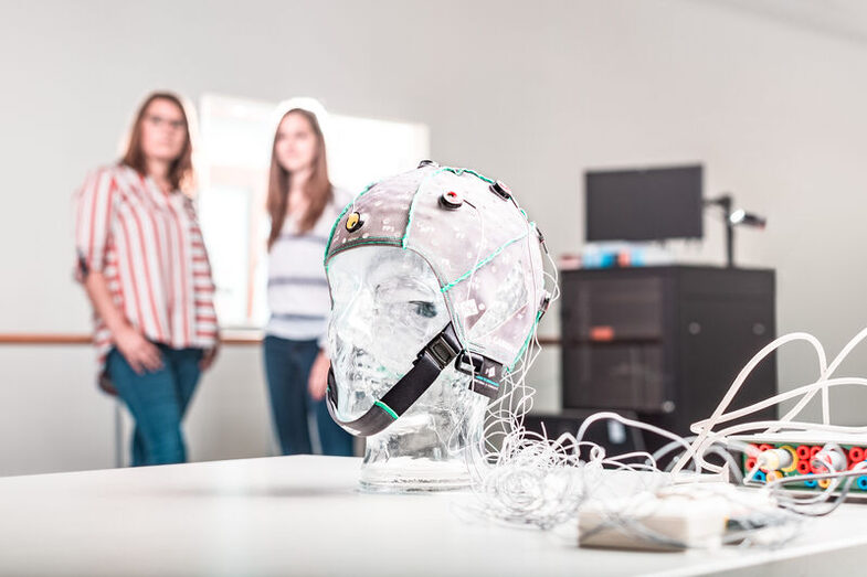 Two women can be seen out of focus in a laboratory, with a transparent head wearing an EGG mask in the foreground.