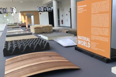 Blick in das Foyer des Fachbereichs Architektur. Auf einem Tisch steht eine kleine Informationstafel zur Ausstellung "Material World". Davor liegen verschiedene Materialproben nebeneinander aufgereiht.