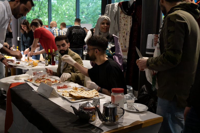 A cultural stand from Palestine is on display. On the table is a sumptuous buffet with various dishes and drinks typical of the country. Behind it is a group of three students who are in charge of the stand.