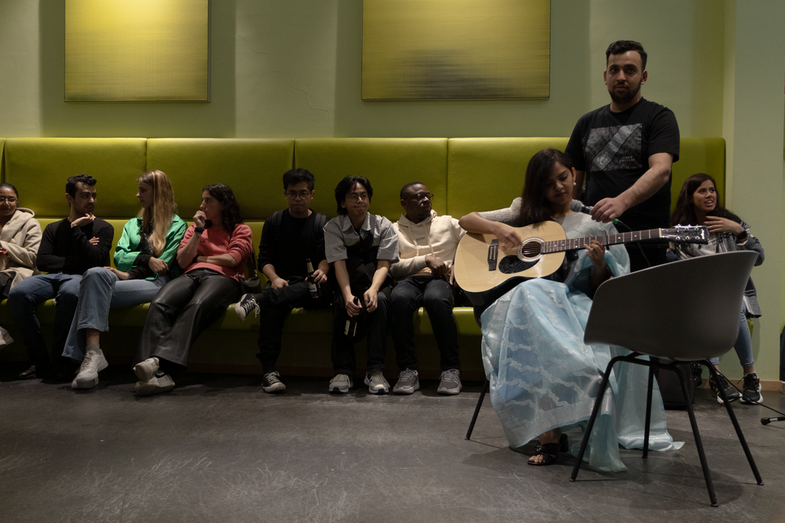 A student with a guitar can be seen in the foreground. She is sitting on a chair and playing a song to which she is singing. A number of students can be seen in the background watching her.