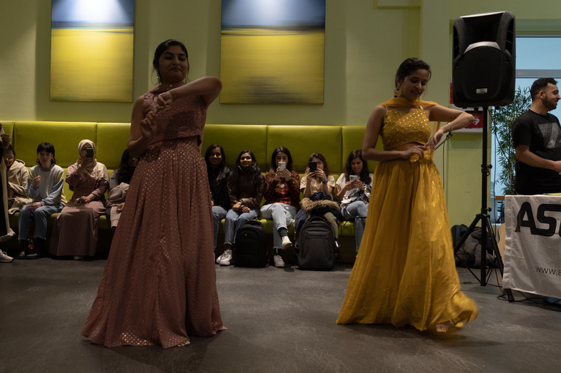 In the foreground, two students in colorful dresses can be seen performing a dance. In the background, a number of students can be seen watching them.