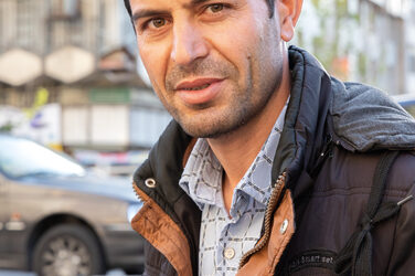 A person in a brown jacket, seen from the head to the elbows, looks into the camera. A street scene can be seen behind her.