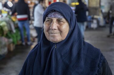 A person is wearing a blue scarf on their upper body and over their head. Her face is friendly, her gaze goes past the camera to the left. A market scene can be seen behind her.