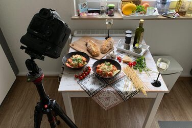 Eine Kamera auf einem Stativ links vorn ist auf einen Tisch gerichtet, darauf zwei Teller mit einem Nudelgericht, umgeben von entsprechenden Zutaten und Gewürzen.