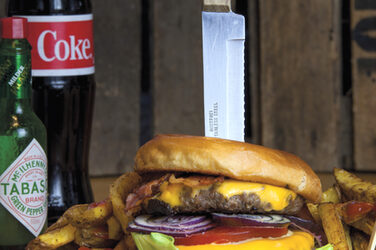 auf einem Couchtisch das Motiv: Ein Burger, in dem senkrecht ein Messer steckt,  auf einem Holzbrett. Dazu eine Cola- und eine Gewürzflasche.