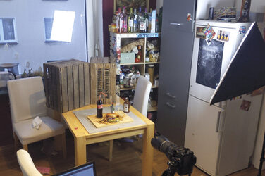 Living room setting for a photo of a burger: in the foreground a camera on a tripod, next to it a laptop on a chair. In the middle ground on a coffee table the motif: a burger with a knife stuck vertically into it on a wooden board. In addition, a bottle of cola and a bottle of spices.