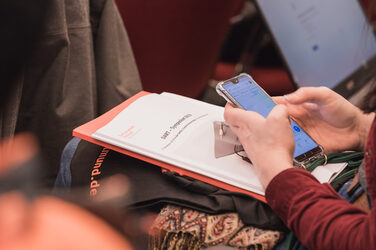 Eine Person blickt auf ihr Smartphone. Auf ihrem Schoß liegen eine Tasche mit FH-Logo, eine orangene Mappe und Unterlagen des DART-Symposiums.