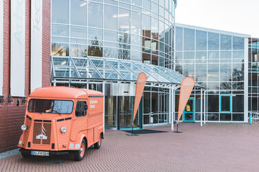Ein Gebäude mit einer Glasfront. Vor dem Gebäude steht der orangene Citroën HY mit dem Logo der Fachhochschule Dortmund.