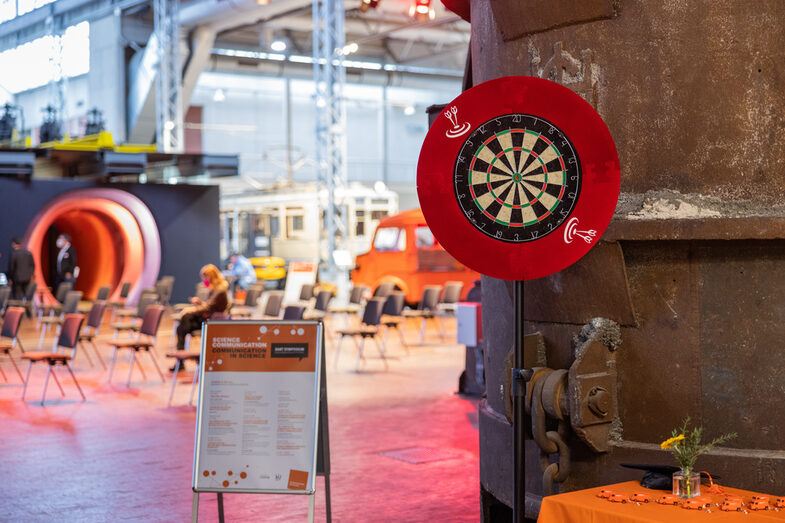 Dart board and the program poster for the DART Symposium 2021 in front of chair rows in a big hall at DASA Working World Exhibition.