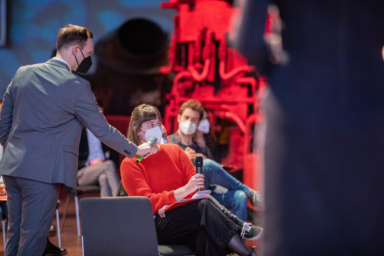 An audience member wearing a mask asks a question into a microphone.