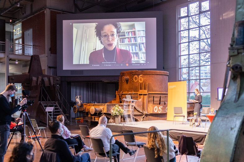 The audience looks up to a  Prof. Dr. Stefanie Molthagen-Schnöring who is connected via video and  projected on a big screen next to the stage.
