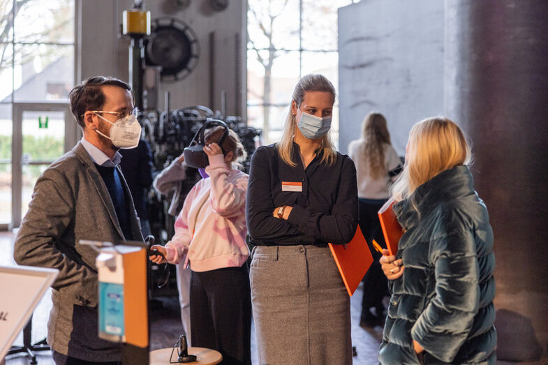 Three members of the jury talk in the exhibition. In the background participants use VR-goggles.