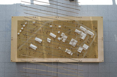 Modellaufsicht mit mehreren eingeschossigen Gebäudemodellen aus weißer Pappe,<br>darüber gespannt eine Bahn gebundener Bambusstäbe.