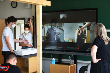 Auf einem großen Bildschirm werden die Erstsemester-Gruppen per Videoübertragung gezeigt. Vor und neben dem Bildschirm arbeiten weitere Personen an ihren Projekten.