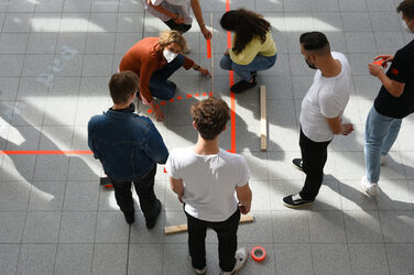 In der Nahaufnahme sieht man von oben, wie Erstsemester den Grundriss ihrer WG mit neon-orangenem Klebeband auf den Boden im Foyer kleben.