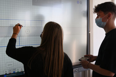 Zwei Studierende stehen vor einem großen Screen. Die Studentin links zeichnet den Grundriss auf den interaktiven Bildschirm. Der Student rechts schaut zu und hält ein Smartphone in der Hand.