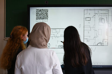 Drei Studentinnen stehen vor einem großen Bildschirm. Die Studentin rechts ergänzt mit einem digitalen Stift den bereits gezeichneten Grundriss auf dem Screen.