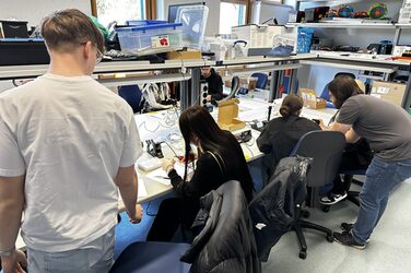 In one room, several people sit and stand at tables, the room is filled with technical objects.