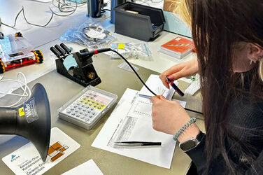 Several technical objects are lying on a table, a person is sitting next to them holding a small pair of pliers with a cable in their hands.