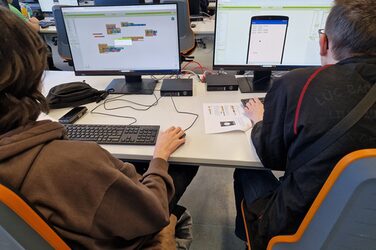 Two people are sitting at a table with two screens.