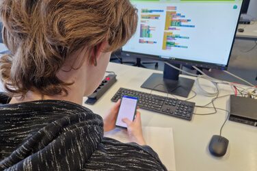 A person sits at a table with a computer and holds a smartphone in front of them.