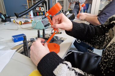 Two hands hold a table lamp, technical devices can be seen in the background.