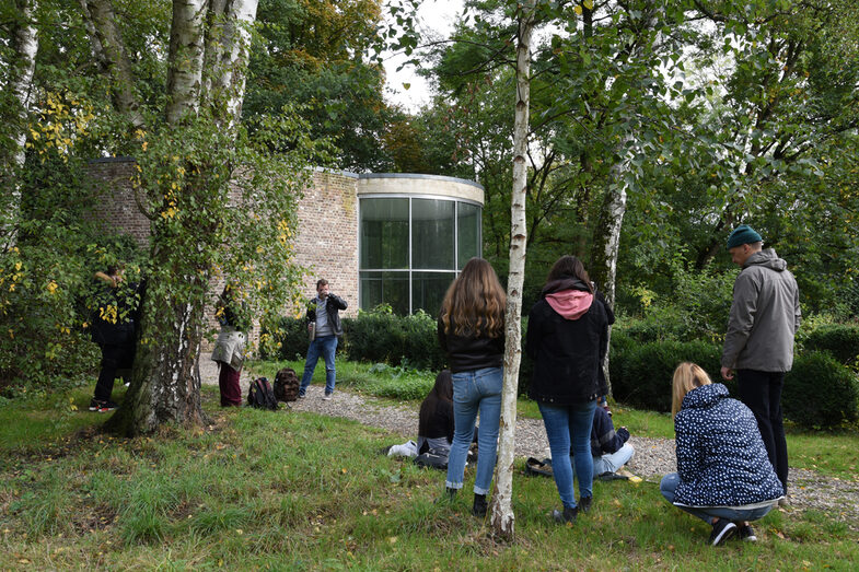 People standing on a gravel path to a building between bushes and trees