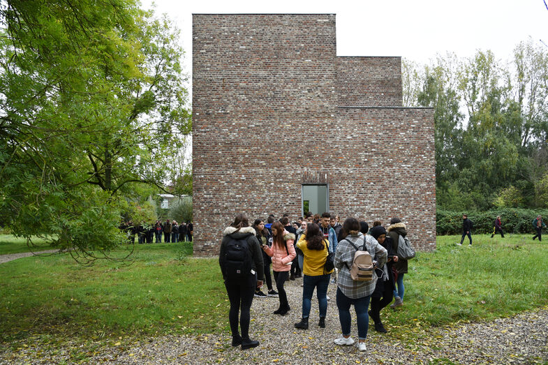 People are standing in front of a building. The building is surrounded by a meadow and trees.