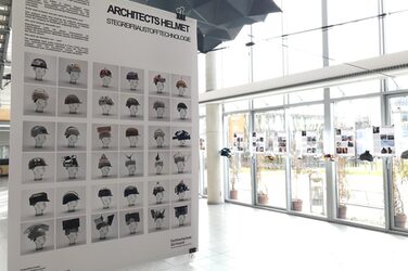 The picture shows an explanatory poster in the foyer of the Faculty of Architecture with the title "ARCHITECTS HELMET, STEGREIF BAUSTOFFTECHNOLOGIE" and depicts 36 differently designed, colorful, plastic and geometrically highly modified construction helmets. Explanatory posters with the corresponding helmets hang in the background. The students' posters explain the key aspects of a given architect's architectural style and the choice of materials. An example building is also presented, which was used as inspiration for the design of the helmet.