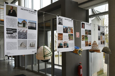 The picture shows four explanatory posters with the corresponding architects' helmets, as they hang in the foyer of the Faculty of Architecture. They are decorated with metal buttons or modeling clay in the style of Otto Wagner or Antoni Gaudí, for example.
