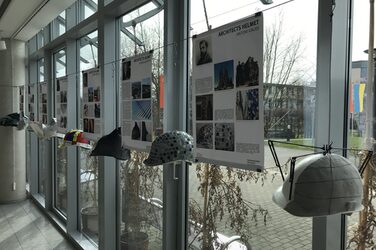 Das Bild zeigt vier erläuternde Plakate mit den dazugehörigen Architekten-Helmen, wie sie im Foyer des Fachbereichs Architektur hängen. Sie sind zum Beispiel in Anlehnung an Günther Behnisch mit Mosaikplättchen bekleidet.