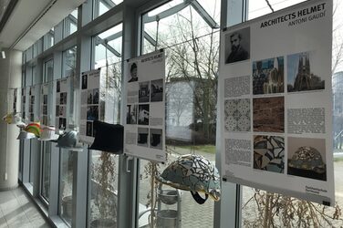 The picture shows four explanatory posters with the corresponding architects' helmets as they hang in the foyer of the Faculty of Architecture. They are wrapped in black fabric in the style of Luigi Snozzi, for example.