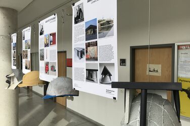 Das Bild zeigt vier erläuternde Plakate mit den dazugehörigen Architekten-Helmen, wie sie im Foyer des Fachbereichs Architektur hängen. Sie sind zum Beispiel in Anlehnung an Ludwig Mies van der Rohe mit Beton bedeckt und einer aufgestelzten quadratischen Plattform versehen.