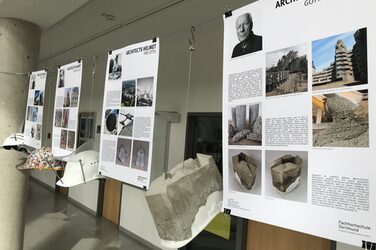 The picture shows four explanatory posters with the corresponding architects' helmets as they hang in the foyer of the Faculty of Architecture. In reference to Gottfried Böhm, for example, they are covered with heavy, massive and angular concrete volumes.