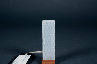 In this picture, a 3D-printed model made of translucent plastic, about three centimetres high, is presented on a thin wooden base. It shows the Hearst Tower in New York City, a tower with a zig-zag silhouette whose surrounding glass façade is composed of large triangular elements.