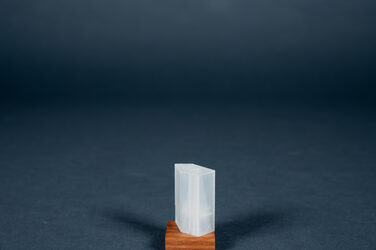 In this picture, a 3D-printed, approximately two-centimeter-high model made of translucent plastic is presented on a thin wooden base. It shows the Bruder Klaus Feld Chapel in the Eifel region, a tower building with a pentagonal basic shape and monolithic walls of varying lengths and heights.