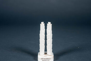 This picture shows a small model made of white plastic the size of a chess piece. The two towers are the same height and stand on two square marble bases with strongly rounded corners and the name of the building as an inscription. The towers are the Marriot Marquis Hotel in Dubai in the United Arab Emirates. They both have a strongly oval basic shape and fan-shaped staggered levels with complex geometry surrounding the building.