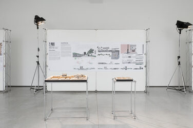 Blick auf die Ausstellungsinszenierung von Studentin Anna Nölle mit den Entwurfsmodellen im Vordergrund und der Ausstellungswand mit den Entwurfsplänen im Hintergrund.