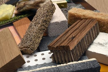 The picture shows a close-up of a disordered collection of different materials in a rectangular shape of the same dimensions with different orientations.
In the foreground of this picture are a solid, brown block of wood, a fibrous, solid piece of brown wood fiber, a composite piece made of a red-brown wooden board with grooves, with a thick piece of cardboard glued to the underside, a gray-black piece of polystyrene, the front of which is plastered white, a white, solid, porous piece of sand-lime brick and a light brown fibrous piece of wood wool.
Underneath are other similar pieces of material of this type.