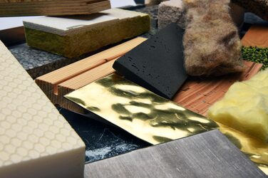 The picture shows a close-up of a disordered collection of different materials in a rectangular shape of the same dimensions with different orientations.
In the foreground of this picture are a beige-colored block of Styrodur with a wavy relief on the surface, a thin, beaten and wavy golden-yellow strip of sheet metal, a thin strip of black foam, a thin, smooth, gray strip of material, a beige-yellow piece of wood with a long notch, a brown-grey-black, somewhat tattered strip of sheep's wool, a piece of composite insulation, i.e. a piece of rock wool with other layers firmly glued on, and a dense, yellow piece of glass wool.
Underneath are other similar pieces of material of this type.