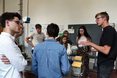 The picture shows a group of young people in the concrete laboratory of the Faculty of Architecture at the Fachhochschule Dortmund. On the right-hand side of the picture, research assistant Mr. Daniel Horn is explaining the concrete mixing process on a small scale to the students spread around a low table. He is holding the metal bowl in which the concrete is freshly mixed. The steel formwork molds can be seen on the table, in which some of the students have already placed their own cut Styrodur formwork for their self-designed sculptures. The orange-colored Styrodur formwork is held in the larger, cubic steel forms by clamping them together from the outside with screws.
