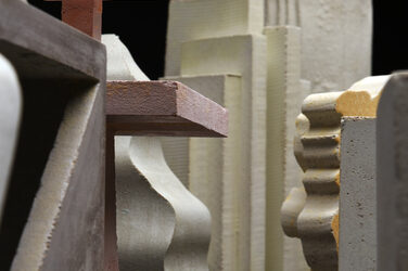 The picture shows a close-up of a collection of concrete sculptures of different dimensions and generally different designs. The figures fill the entire picture.
In the foreground of this picture is a cubic sculpture with a triangular recess on the left edge of the picture, in the middle background on the left is a figure that looks like a pyramid with undulating surfaces, on the right is a bar-shaped figure with strongly varying undulating side surfaces, in the background in the middle of the picture is a bar-shaped figure consisting of about six rectangles that are staggered in height towards the sides.
There are other concrete sculptures of this type in the surrounding area.