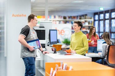 Photo of a library employee behind the counter. She is in conversation with the student opposite her who has books in hand. __ The female employee stands behind the counter of the library and is in conversation with the student opposite her who has books in hand.