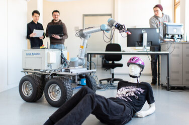 Foto eines Rettungsroboter, der neben einer lebensgroße Puppe auf dem Boden steht. Dahinter links sind zwei Männer mit Unterlagen, die zum Roboter schauen. Rechts hinten ist ein Mann, der an einem Tisch steht und in den Rechner schaut.