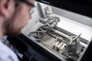 Photo of a man using a brush to remove excess gray powder from a printed component in a glove box.