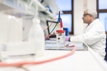 Photo of a test setup in the corrosion laboratory of the Faculty of Mechanical Engineering. Behind it, an employee is sitting at a computer in a blur. The man is wearing a white coat and protective goggles.