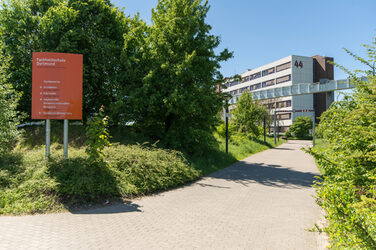 Foto vom Gehweg, der zum Gebäude Emil-Figge-STraße 44 der Fachhochschule Dortmund hinführt. Rechts neben dem Gehweg ein Schild als Wegweiser mit dem Logo der Fachhochschule Dortmund.