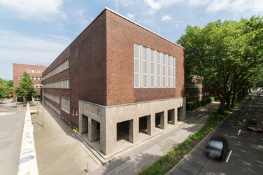 Photo with a view of parts of a building of the Fachhochschule Dortmund at Max-Ophüls-Platz with forecourt and sidewalk as well as street with cars next to it.