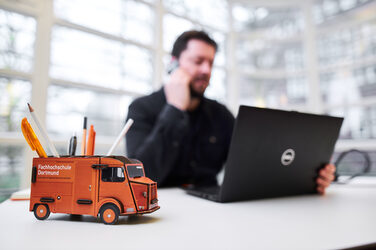 Foto einer Stiftebox in Form des Kastenwagens der Fachhochschule auf einem Schreibtisch. Unscharf dahinter sitzt ein Mann am Laptop und telefoniert.__Photo of a pencil box in the form of a car on a desk. Out of focus behind it, a man sits at his laptop and makes a phone call.