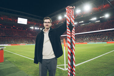 Eine Person steht am Spielfeldrand in einem Stadion und präsentiert einen Fanschal.